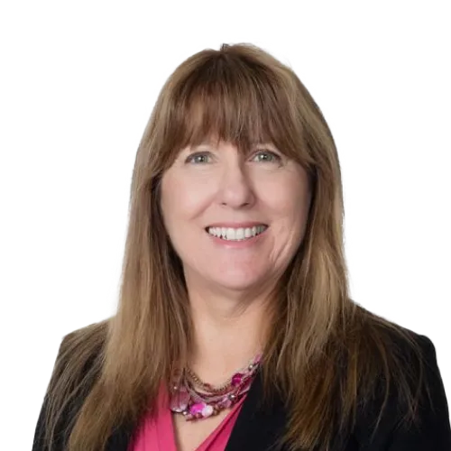 A woman with long light brown hair and bangs smiles at the camera. She is wearing a black blazer over a pink top and a pink beaded necklace. The background is plain white.
