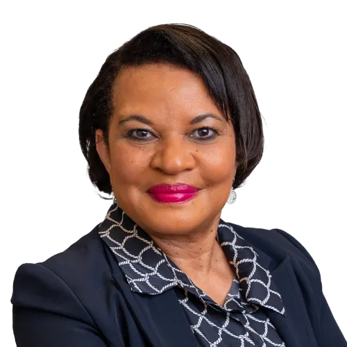 A woman with short, dark hair and medium brown skin smiles at the camera. She wears a dark blazer over a patterned blouse with a chain-link design. She has bright pink lipstick and subtle eye makeup. The background is white.