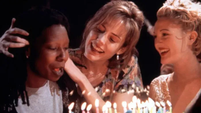 Three women sit close together, smiling and celebrating in front of a brightly lit birthday cake. One woman is blowing out the candles while the others look on joyfully, their faces illuminated by the candlelight. The mood is warm and festive.