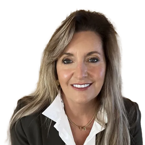 A woman with long blonde hair smiles at the camera. She is wearing a dark blazer over a white collared shirt and a gold necklace. The background is transparent.