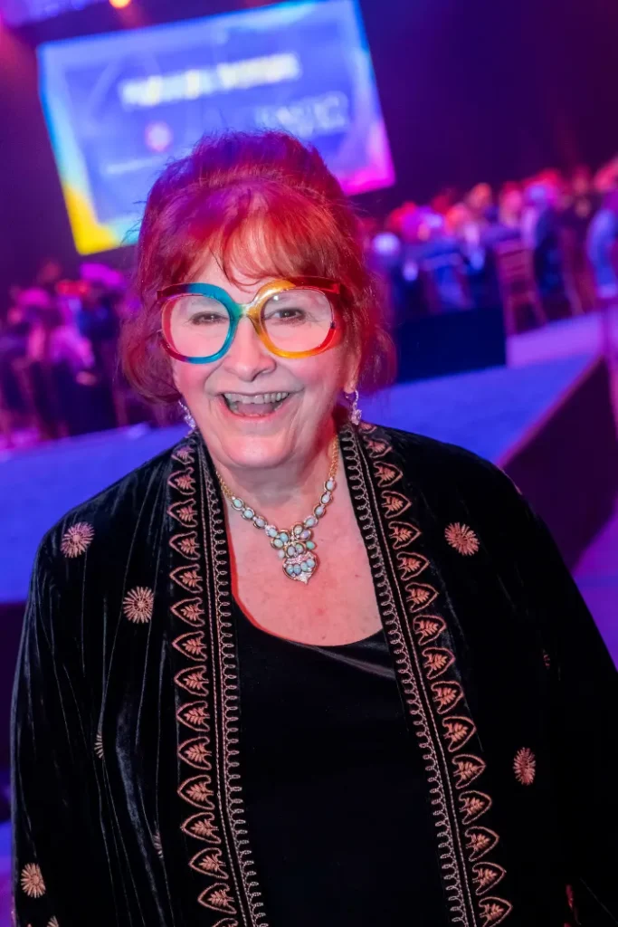 An older woman with red hair and rainbow-rimmed glasses smiles at an indoor event, where $2.3 Million Raised at 30th Annual The Chase celebrated DAP Health’s 40th Anniversary amid colorful lights and a vibrant crowd.