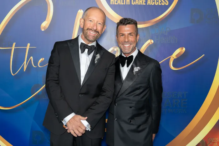 Two men in black tuxedos and bow ties stand smiling before a blue and gold backdrop highlighting $2.3 Million Raised at 30th Annual The Chase Celebrating DAP Health’s 40th Anniversary, featuring decorative script and event branding.