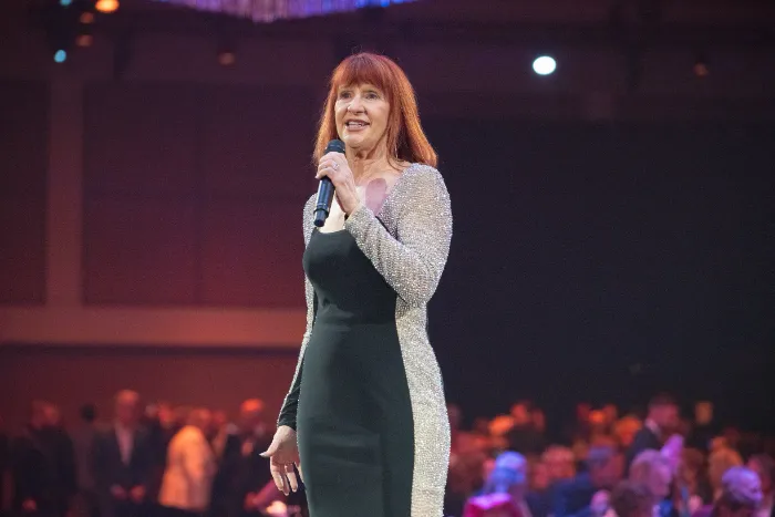 A woman with straight red hair stands on stage holding a microphone, wearing a black dress with glittery silver sleeves and sides. She smiles at the audience during the $2.3 Million Raised at 30th Annual The Chase Celebrating DAP Health’s 40th Anniversary.