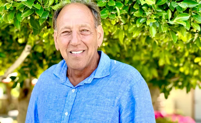 A smiling middle-aged man with short, graying hair wears a light blue shirt and stands outdoors in front of lush green foliage, with sunlight filtering through the leaves and creating a bright, cheerful atmosphere.
