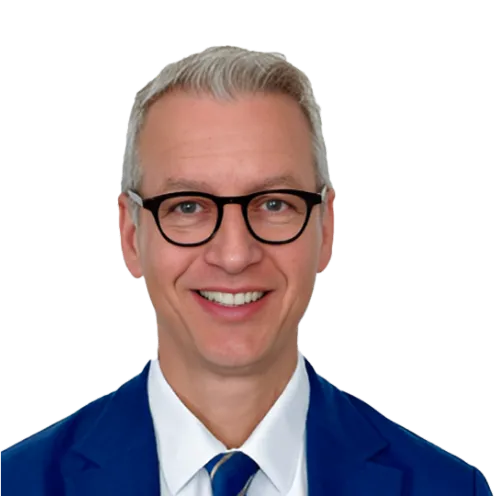 A smiling man with short, neatly combed gray hair, wearing black-rimmed glasses, a white shirt, a blue suit jacket, and a blue striped tie, facing forward against a transparent background.