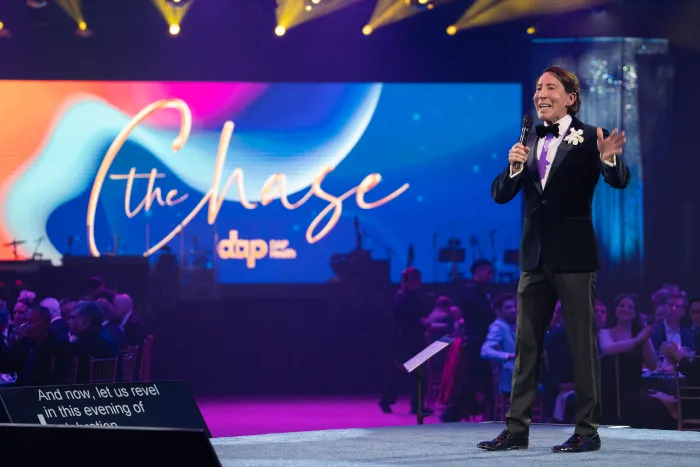 A man in a tuxedo and bow tie stands on stage holding a microphone, addressing an audience. Behind him, a screen displays "The Chase: $2.3 Million Raised at 30th Annual, Celebrating DAP Health’s 40th Anniversary." Stage lights create a festive atmosphere.