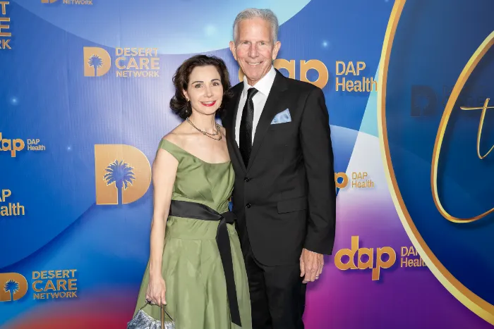 A smiling woman in a green off-shoulder dress stands arm-in-arm with a man in a black suit before a blue and purple step-and-repeat backdrop at The Chase, where $2.3 Million was raised celebrating DAP Health’s 40th Anniversary.