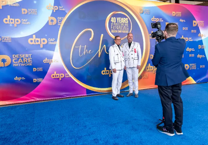 Two men in matching white suits with black trim pose on a blue carpet at the $2.3 Million Raised at 30th Annual The Chase Celebrating DAP Health’s 40th Anniversary, against a colorful logo-filled backdrop. A photographer captures the moment.