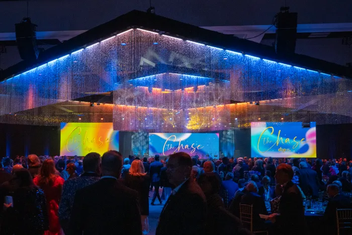 A large crowd attends a glamorous indoor event under shimmering lights, as two screens display “The Chase.” $2.3 Million Raised at 30th Annual The Chase Celebrating DAP Health’s 40th Anniversary adds excitement to the elegant, festive atmosphere.