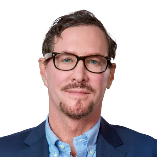 A middle-aged man with short brown hair, light skin, and glasses. He has a trimmed goatee and mustache. He is wearing a blue button-up shirt under a dark blazer, looking directly at the camera with a neutral expression.