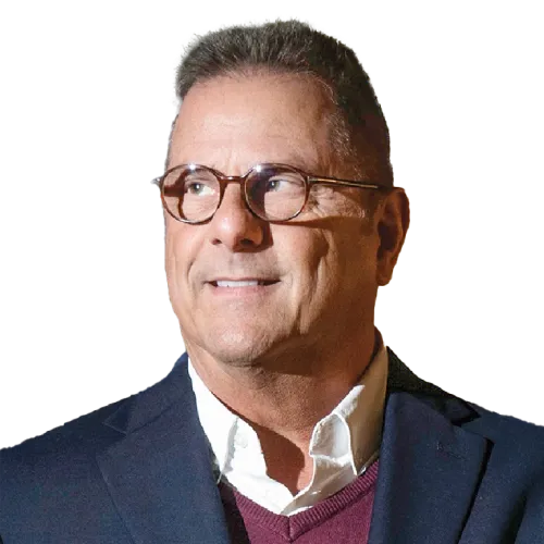A middle-aged man with short brown hair and glasses looks to his right, smiling slightly. He wears a dark blazer over a maroon sweater and white collared shirt. The background is plain and light-colored.
