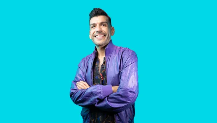 A smiling man with short dark hair stands against a solid turquoise background. Wearing a shiny purple jacket over a patterned shirt, he has his arms crossed, giving an approachable and cheerful vibe—perfect for celebrating at a Pride Parade.