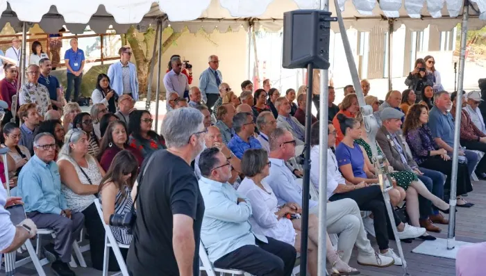 A diverse group of people sit closely together on white folding chairs under a tent at an outdoor event, attentively listening. Some stand or take photos. Most appear engaged, with a few talking quietly, as sunlight filters through the tent.