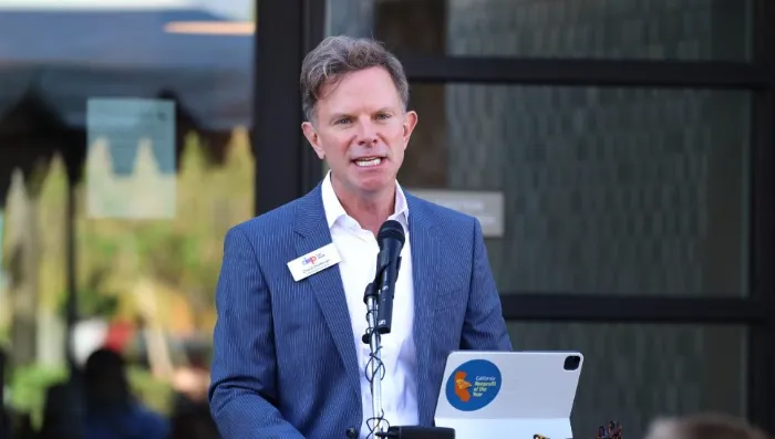 A man in a blue suit speaks at an outdoor podium with a microphone. He wears a white shirt, no tie, and a name tag. A laptop with a colorful sticker sits on the podium. The background is blurred with plants and a building visible.