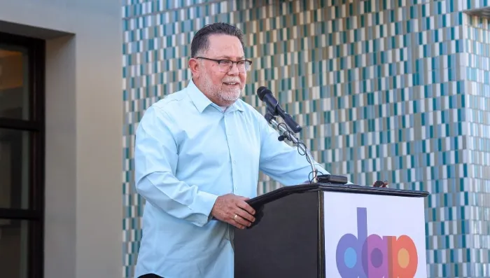 A man with short dark hair and glasses speaks at a podium with a microphone. He is wearing a light blue shirt. Behind him is a modern-looking, tiled wall. The podium displays the letters "dap" in colorful lowercase font.