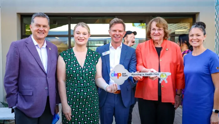 Five smiling adults stand together outside a building. The central person holds a large ceremonial key with "Housing is Health Care" written on it. They are dressed in business or smart-casual attire, posing for a group photo at an event.