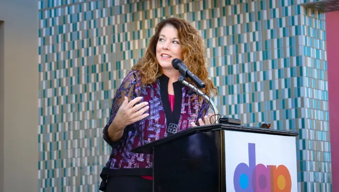 A woman with long curly brown hair speaks at a podium with a microphone. She wears a purple-patterned jacket and gestures with her left hand. The podium displays a colorful "dap" logo. The background features a blue and white tiled wall.