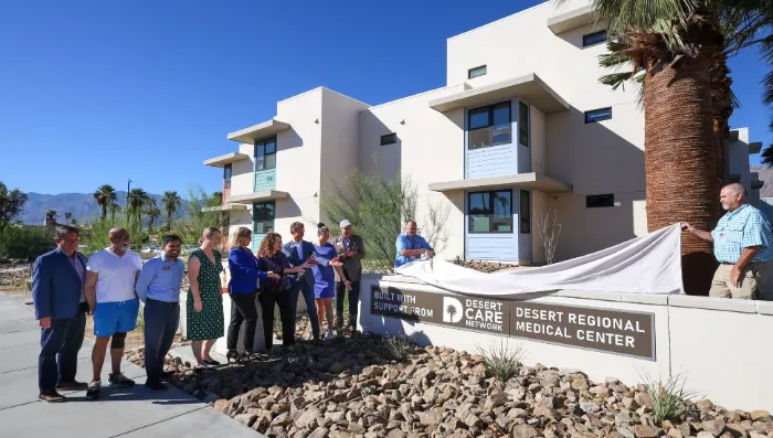 A group of people stand outside a modern beige building, some holding a large white cloth being pulled off a new sign that reads, “Desert Care Network Desert Regional Medical Center.” Palm trees and rocks are visible in the landscaped area nearby.
