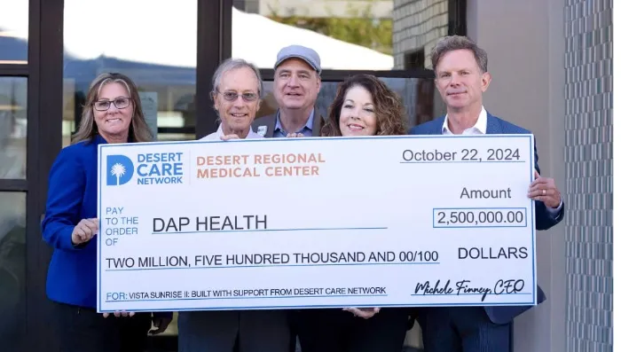 Five people stand smiling and holding a large ceremonial check for $2,500,000 made out to DAP Health from Desert Care Network and Desert Regional Medical Center, dated October 22, 2024. The group poses outdoors in business attire.