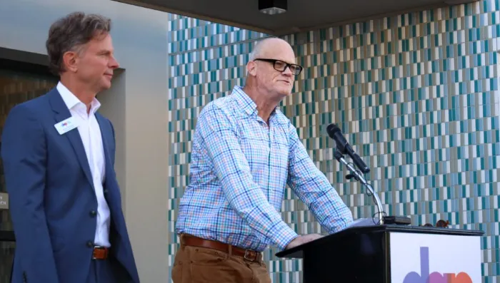 Two men stand outside by a podium. One, in a blue plaid shirt and glasses, speaks into a microphone. The other, in a blue suit, stands nearby. Behind them is a blue, white, and teal tiled wall. The podium has a colorful, partially visible logo.