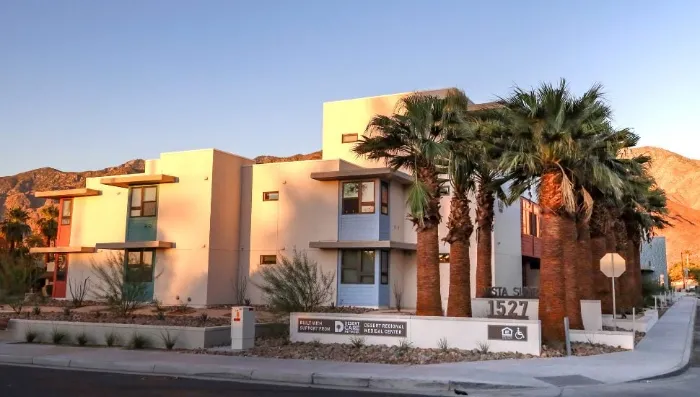Modern apartment complex with a mix of tan, white, and brown facades, large windows, and balconies. Palm trees and desert landscaping surround the building. Mountains and a clear blue sky are in the background. Address sign reads “1527.”.