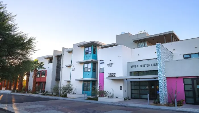 Modern, multi-story building with white walls, teal and pink accents, labeled “David Gluckstein Building 1571.” Large windows, desert landscaping, palm trees, and clear blue sky are visible. Shadows suggest early morning or late afternoon sunlight.