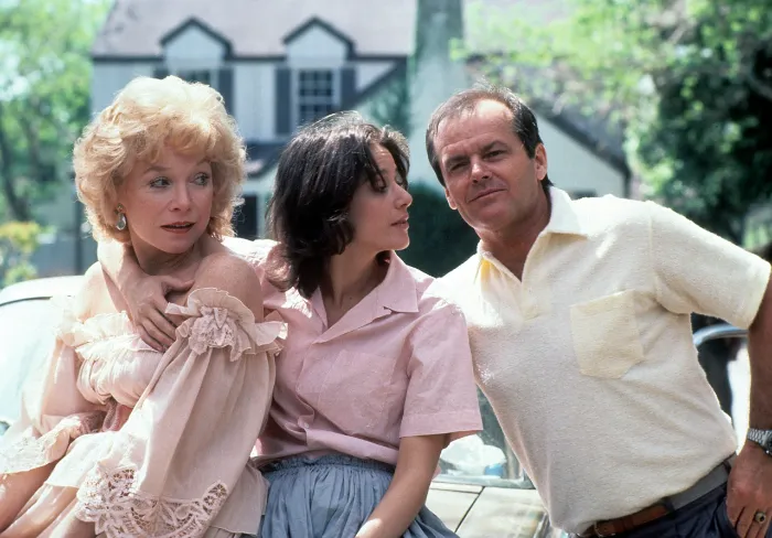 Three people are outdoors in daylight, posing closely in front of a car and a large house. Two women, one older in a frilly dress and one younger in a pink shirt, sit beside a man in a light polo shirt. All are looking in different directions.