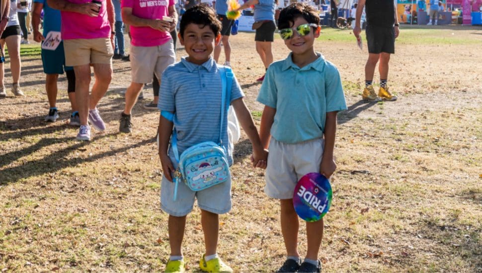 Two young boys stand smiling and holding hands outdoors on dry grass. Both wear light blue shirts and shorts. One has a blue backpack, and the other holds a rainbow “Pride” hat and wears sunglasses. People and colorful booths are visible in the background.