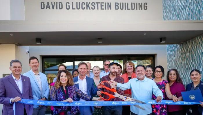 A group of people stands smiling in front of the "David Gluckstein Building" entrance. The central figure holds large ceremonial scissors, cutting a blue ribbon. Others gather closely, celebrating the ribbon-cutting event.