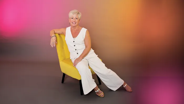 A smiling woman with short blonde hair sits sideways on a yellow chair, wearing a sleeveless white jumpsuit and sandals. The background features a soft gradient of pink, orange, and brown tones. She appears confident and relaxed.