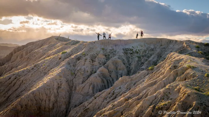 Five people stand with arms raised on a rugged, sunlit desert ridge under a dramatic cloudy sky. The rocky landscape is textured with ridges and shadows. Light streams through the clouds in the background, creating a sense of adventure and vastness.