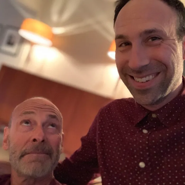 Two men pose indoors. The man on the left, bald with a beard, looks up at the taller man on the right, who has short dark hair and is smiling. Both wear maroon shirts. Warm lighting and two orange lamps are visible in the background.