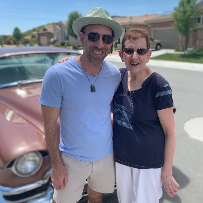 A smiling man in a light green hat, sunglasses, and light blue shirt stands with his arm around a smiling older woman in a navy shirt and glasses. They pose in front of a vintage brown car on a sunny suburban street.