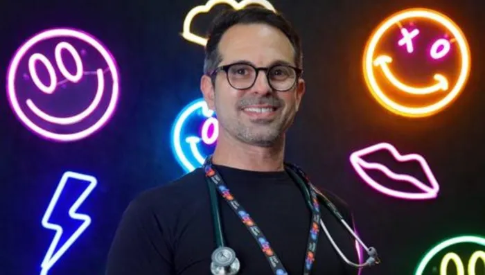 A smiling man with glasses and a stethoscope around his neck stands in front of a black background decorated with colorful neon signs, including smiley faces, a lightning bolt, lips, and a cloud. He wears a black shirt and a patterned lanyard.