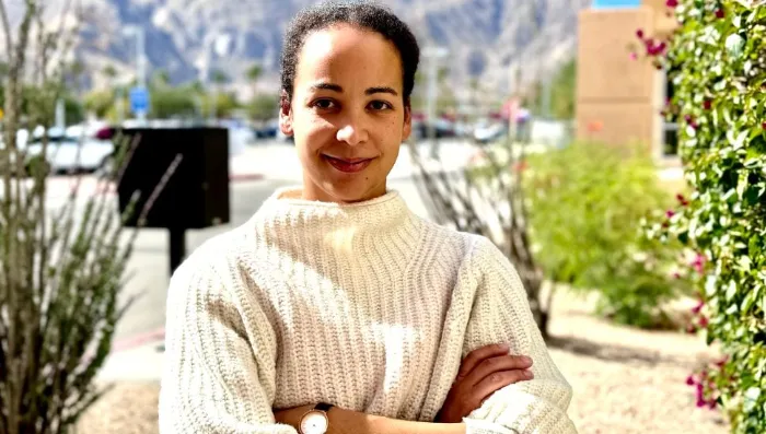 A woman with dark hair tied back, wearing a cream-colored knit sweater and a watch, stands outdoors with arms crossed, smiling softly. The background shows blurred greenery, a building, and distant mountains under a bright sky.
