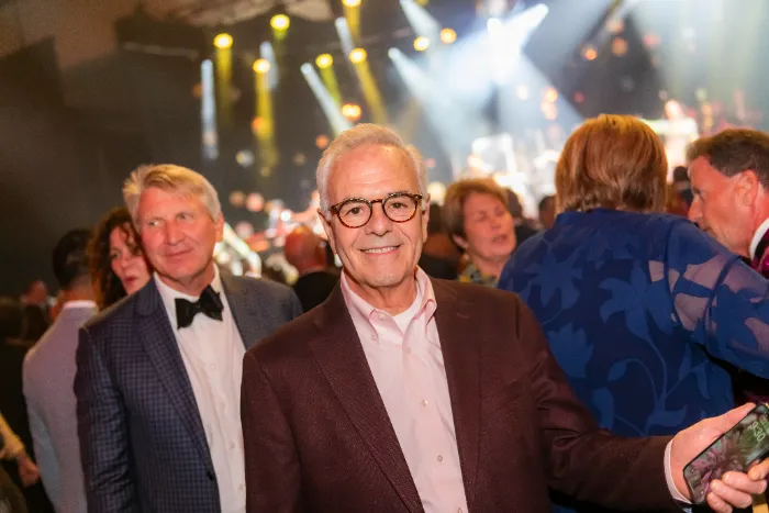 A smiling older man in glasses and a burgundy suit stands at the lively $2.3 Million Raised at 30th Annual The Chase, celebrating DAP Health’s 40th Anniversary. Warm stage lights and formally dressed guests create a festive, energetic atmosphere.