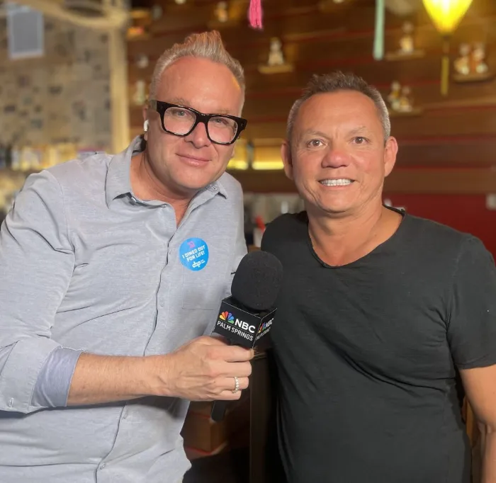 Two men pose indoors. The man on the left wears glasses, a light blue shirt, and holds an NBC microphone, with a "Diner Guy" sticker on his chest. The man on the right wears a black shirt. Both are smiling, with a blurred restaurant background behind them.