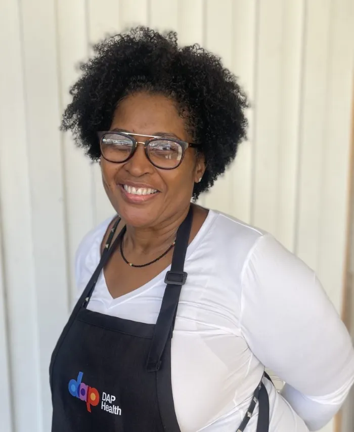 A smiling woman with curly black hair and glasses wears a white shirt and a black apron that says "dap DAP Health" in colorful letters. She stands in front of a white wooden wall, looking directly at the camera.