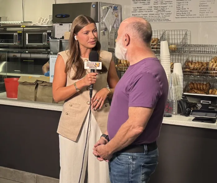 A female reporter in a beige vest and white pants interviews a man in a purple shirt and jeans inside a bakery. She holds a microphone labeled "NEWS 3". Behind them are shelves with bagels, a menu, and kitchen equipment.