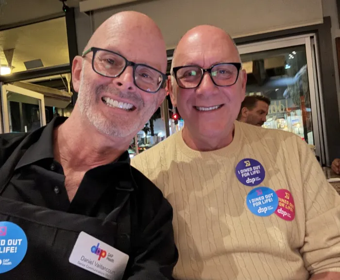 Two smiling older men sit together indoors. One wears a black shirt and nametag, the other a cream sweater with several colorful "I Stand Out For Life!" buttons. Both wear glasses. The background shows a busy restaurant with people and lights.