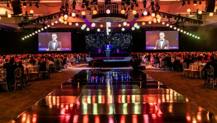 A formal event in a large ballroom features a stage with a speaker in a suit. Two large screens display the $2.3 Million Raised at 30th Annual The Chase, as guests celebrate DAP Health’s 40th Anniversary amid lights and polished floors.