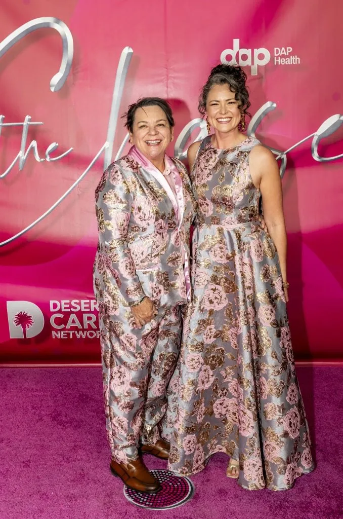 Two people stand smiling on a magenta carpet before a pink backdrop at the 30th Annual The Chase, where $2.3 million was raised celebrating DAP Health’s 40th Anniversary. Both wear matching pink, silver, and gold floral-patterned outfits. Event logos are visible.