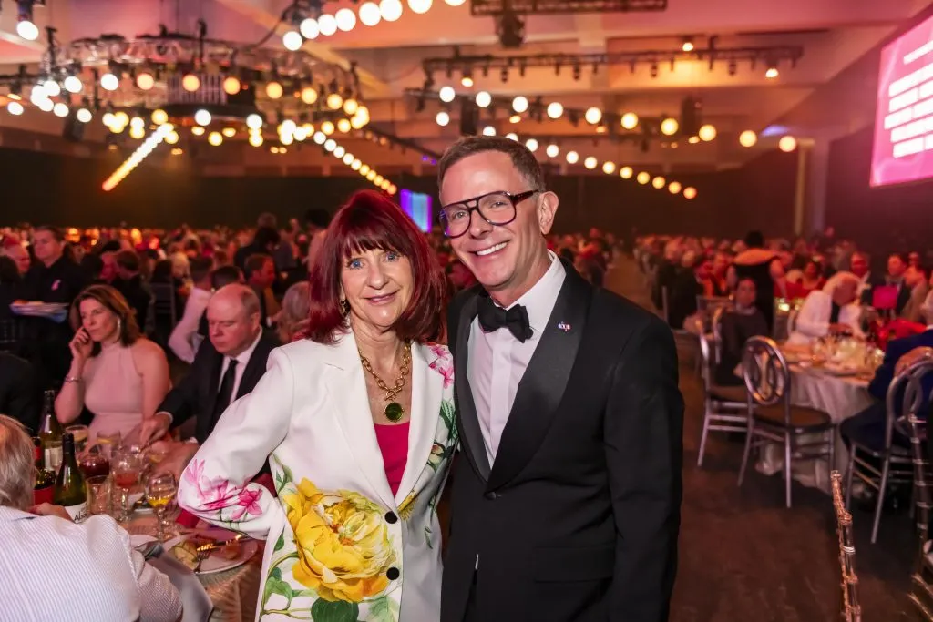 A woman in a white blazer with yellow floral designs stands smiling beside a man in a black tuxedo at the formal $2.3 Million Raised at 30th Annual The Chase, celebrating DAP Health’s 40th Anniversary, with elegant tables and warm lighting in the background.