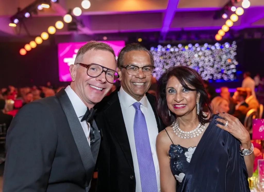 Three adults in formal attire smile at a gala event celebrating DAP Health’s 40th Anniversary. Set against colorful lights and a sparkling stage, the $2.3 Million Raised at 30th Annual The Chase highlights the night’s success.
