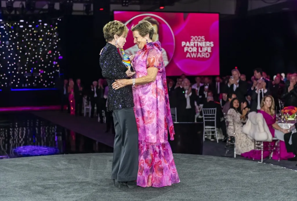 Two older women stand smiling and embracing on a round stage at a formal event celebrating DAP Health’s 40th Anniversary, where $2.3 Million was raised at the 30th Annual The Chase. An audience applauds as they accept the “2025 Partners for Life Award.”.