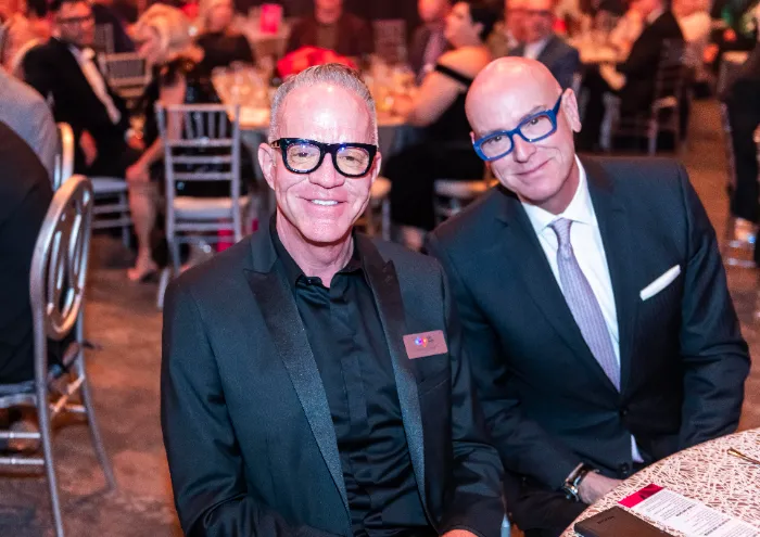 Two men in suits and black glasses sit at a round table at a formal event celebrating $2.3 Million Raised at the 30th Annual The Chase for DAP Health’s 40th Anniversary, with blurred guests and silver chairs filling the background.