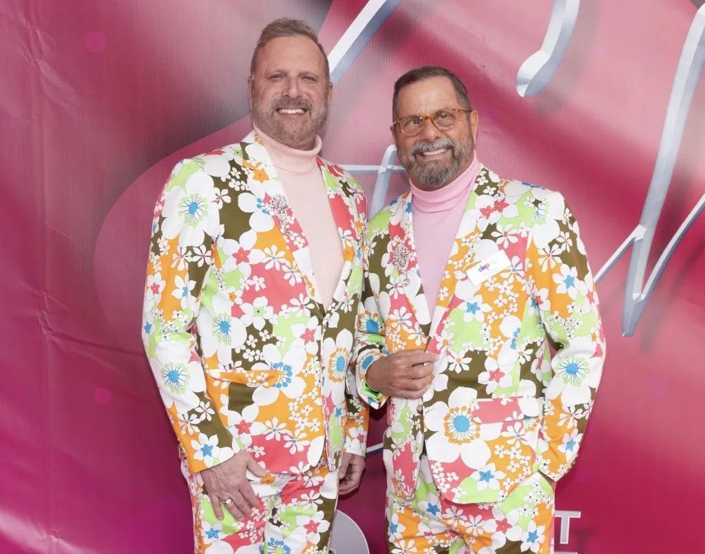 Two smiling men, celebrating $2.3 Million Raised at 30th Annual The Chase honoring DAP Health’s 40th Anniversary, stand in front of a pink backdrop in matching floral suits and pink turtlenecks, arms around each other as they pose for the camera.