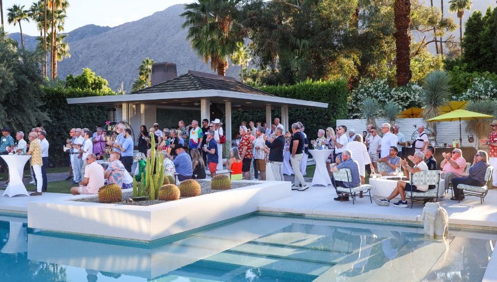 A large group of people socializes at a poolside party with modern patio furniture, desert plants, and palm trees. Mountains are visible in the background. Guests wear colorful, casual clothes, and the sunny setting suggests a warm, festive atmosphere.