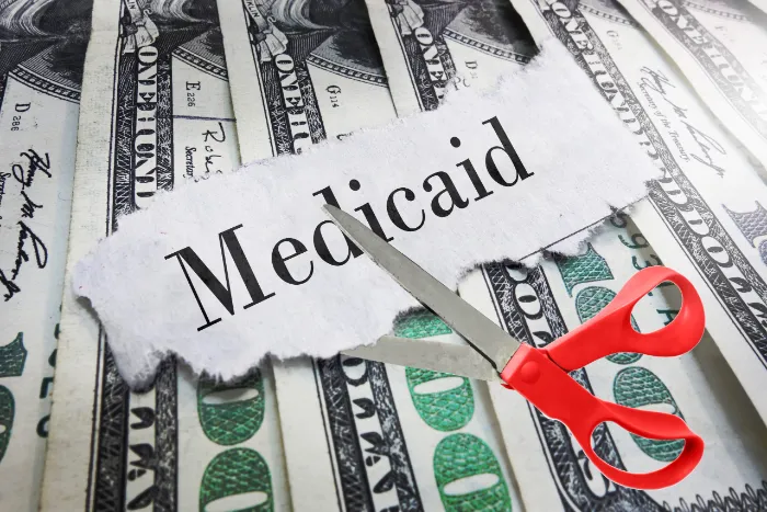 A pair of red scissors cuts through a piece of paper labeled “Medicaid,” placed over several U.S. hundred dollar bills, symbolizing budget cuts or funding reductions for the Medicaid program.