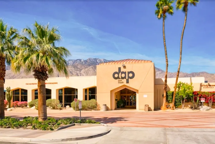 A modern tan building labeled “dap” with large windows, palm trees, and desert landscaping offers Health Services, set against mountains under a clear blue sky. The sunlit entrance is inviting, and the surroundings are tidy.